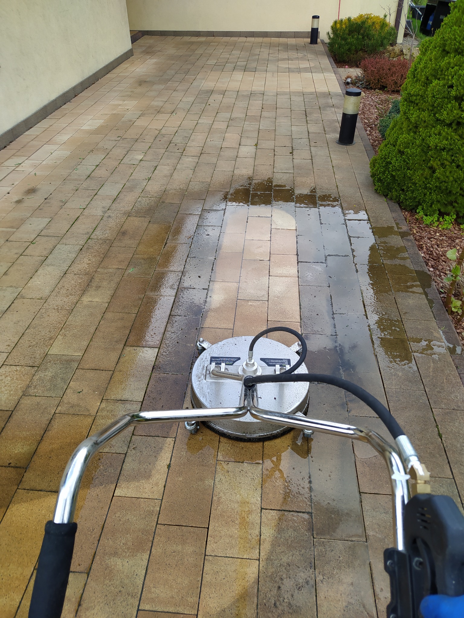 Czyszczenie kostki brukowej na podjeździe za pomocą myjki ciśnieniowej z okrągłą dyszą, widoczne mokre i czyste fragmenty kostki.