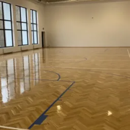 Sala gimnastyczna z parkietem ułożonym w jodełkę, świeżo po cyklinowaniu, z widocznymi liniami boiska i dużymi oknami z czarnymi ramami.