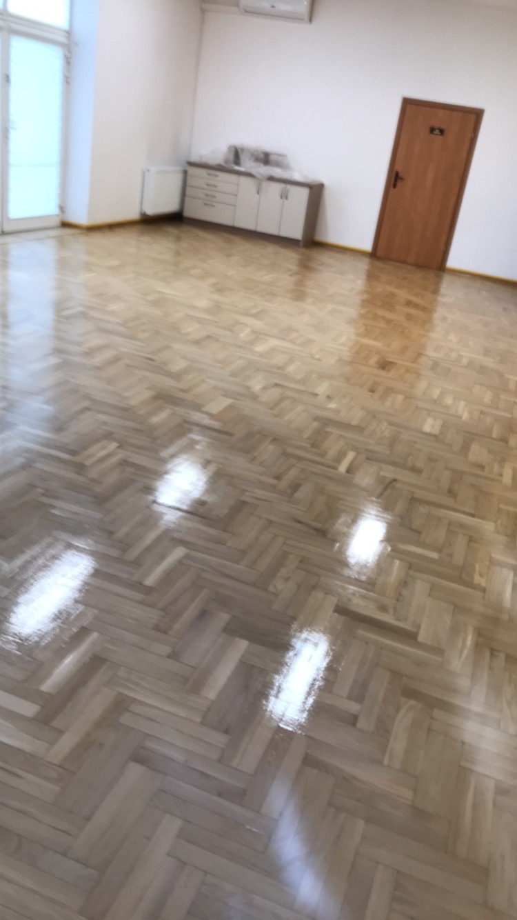 Świeżo polakierowany parkiet ułożony w jodełkę, odbijający światło w pustym pomieszczeniu z białymi ścianami, szafką i brązowymi drzwiami.