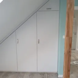 Biała szafa wnękowa z czterema drzwiami, dopasowana do skosu poddasza, z drewnianą belką na pierwszym planie i schodami drabinkowymi w tle.