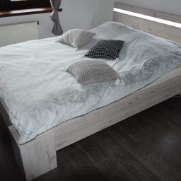 MD-MEBLE.COM - Łóżko z jasnego drewna z podświetlanym zagłówkiem, przykryte puszystą, błękitną narzutą i trzema dekoracyjnymi poduszkami w odcieniach szarości i czerni, na ciemnej podłodze z paneli.
