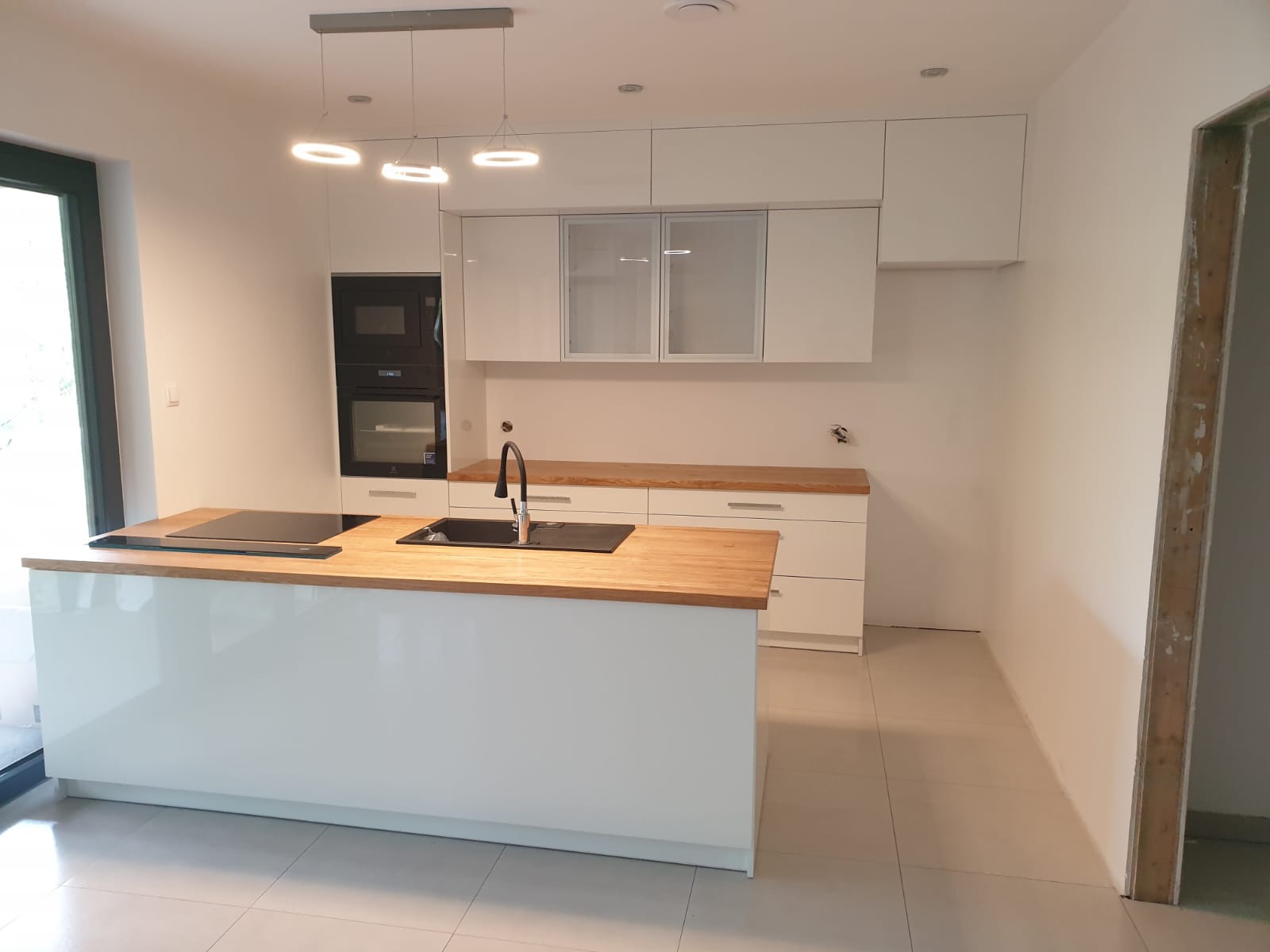 Nowoczesna, biała kuchnia z drewnianym blatem i wyspą kuchenną; widoczne wiszące lampy pierścieniowe i zabudowany piekarnik.