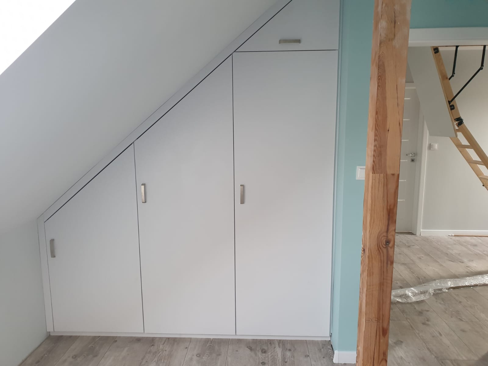 Biała szafa wnękowa z czterema drzwiami, dopasowana do skosu poddasza, z drewnianą belką na pierwszym planie i schodami drabinkowymi w tle.