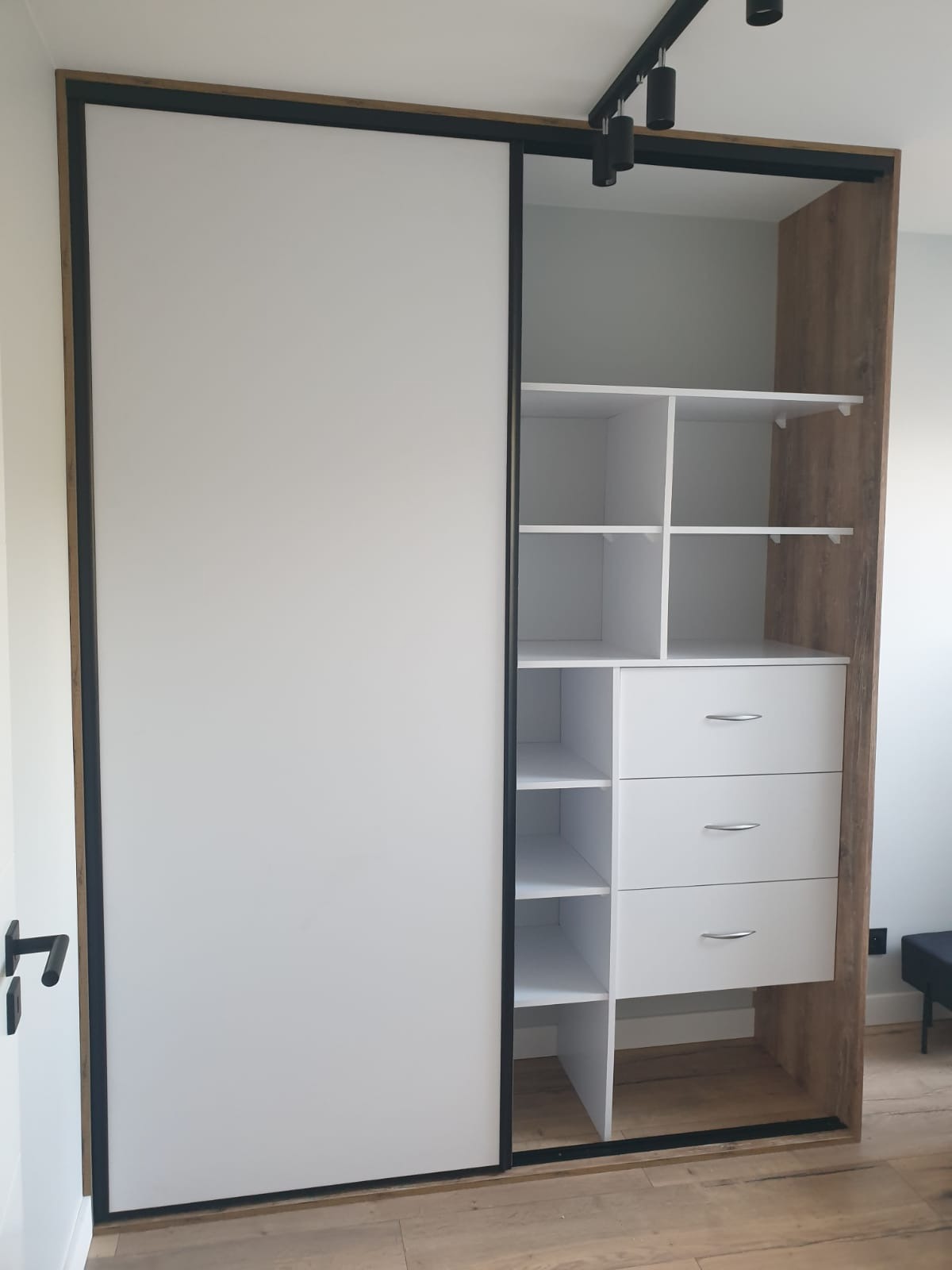 Biała szafa wnękowa z przesuwnymi drzwiami i drewnianym obramowaniem, widoczne półki i szuflady, oświetlenie sufitowe