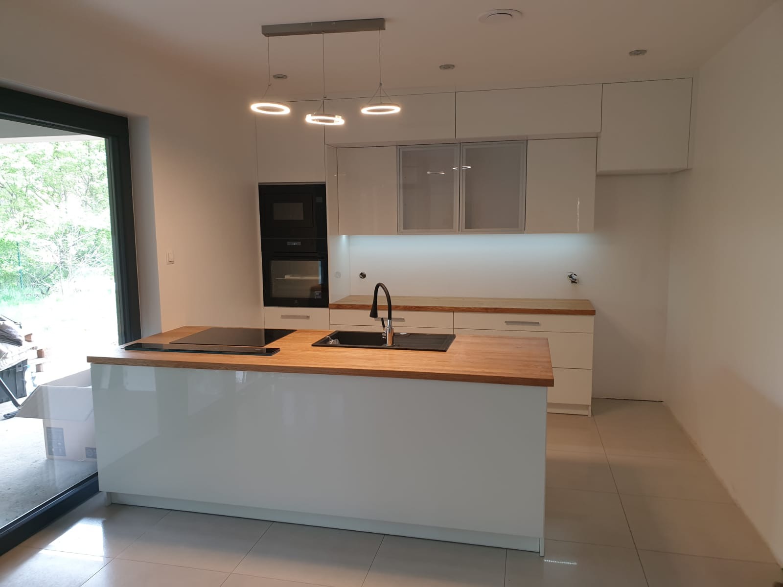 Biała kuchnia z wyspą i drewnianym blatem, nowoczesne oświetlenie wiszące, widoczne okno z widokiem na zieleń.