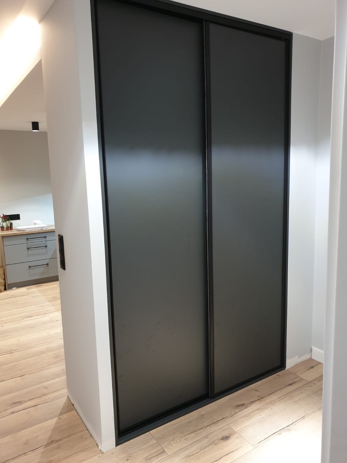 Nowoczesna szafa wnękowa z przesuwnymi, grafitowymi drzwiami i czarną ramą, wkomponowana w jasne wnętrze z drewnianą podłogą.