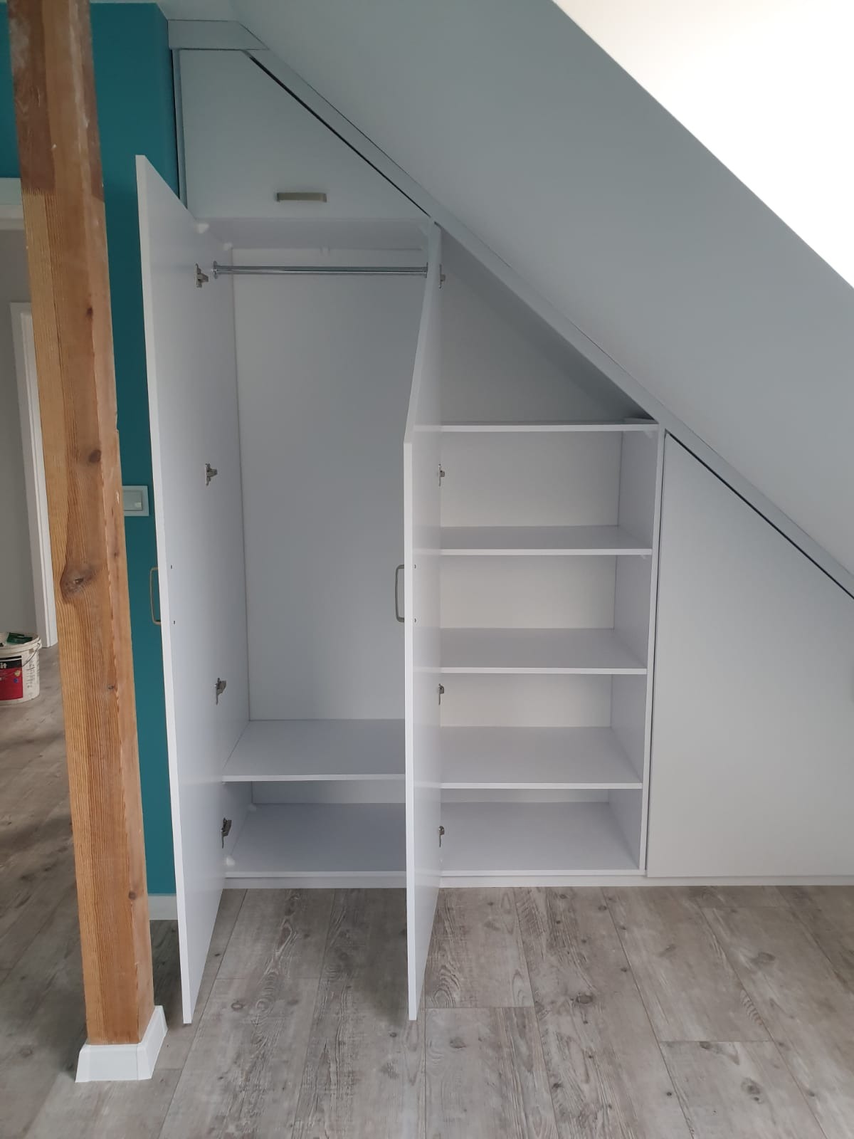 Biała szafa wnękowa z półkami i drążkiem na ubrania, dopasowana do skosu dachu, z otwartymi drzwiami i widocznym drewnianym słupem w tle.