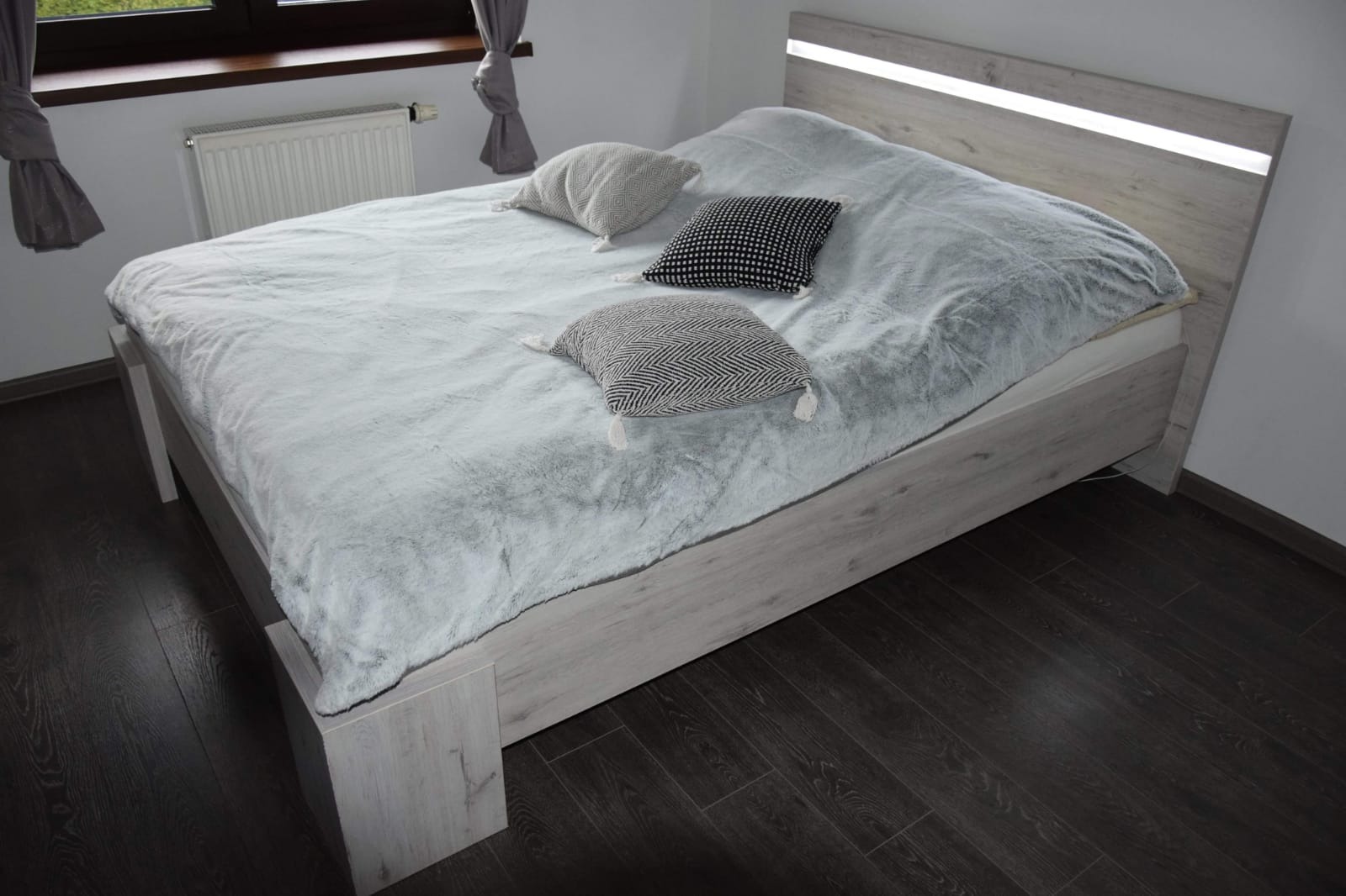 Łóżko z jasnego drewna z podświetlanym zagłówkiem, przykryte puszystą, błękitną narzutą i trzema dekoracyjnymi poduszkami w odcieniach szarości i czerni, na ciemnej podłodze z paneli.