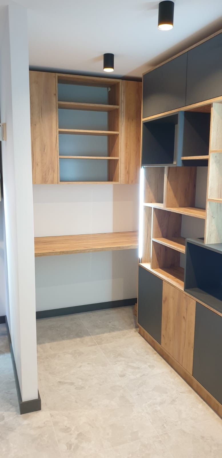 Nowoczesna zabudowa wnęki z biurkiem, szafkami wiszącymi i regałem z podświetleniem LED, fronty w odcieniach drewna i grafitu.