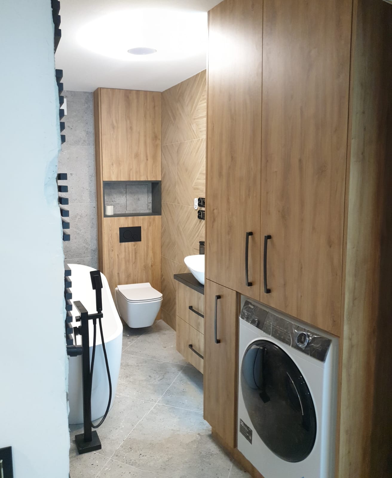 Drewnopodobne szafy na wymiar w łazience z pralką, umywalką i toaletą, szare płytki na podłodze i ścianach, czarne uchwyty i armatura.