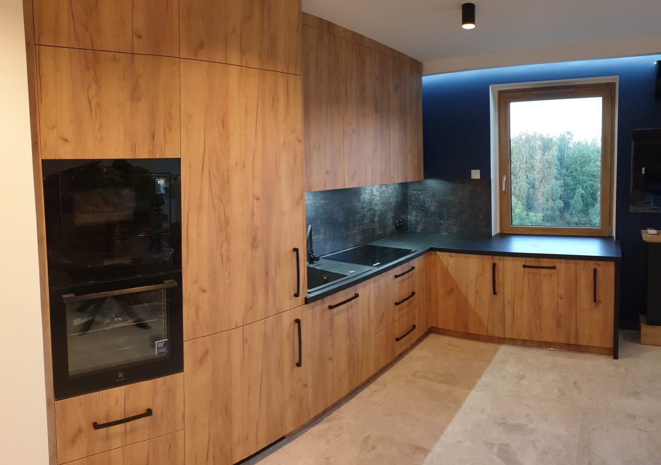 Nowoczesna kuchnia z drewnopodobnymi frontami szafek, czarnym blatem i czarnymi uchwytami, oświetlona punktowo, z widokiem na zieleń za oknem.