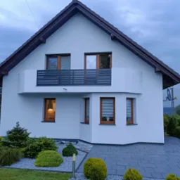 Nowoczesny dom z białymi ścianami, ciemnobrązowym dachem i stalową balustradą balkonową z poziomymi szczeblami, widok z zewnątrz, z zadbanym ogrodem z krzewami i szarym brukowanym podjazdem.