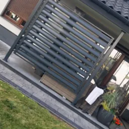 Nowoczesna, szara konstrukcja z poziomych profili, prawdopodobnie zadaszenie tarasu, w Korytnicy. W tle widoczny dom z oknem i roletami zewnętrznymi.