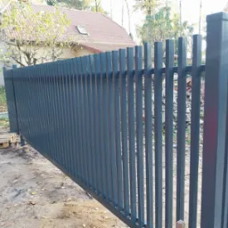 Nowoczesna, przesuwna brama wjazdowa w kolorze grafitowym, z pionowymi profilami, widoczna na tle domu z czerwoną dach&oacute;wką i paletami na ziemi.