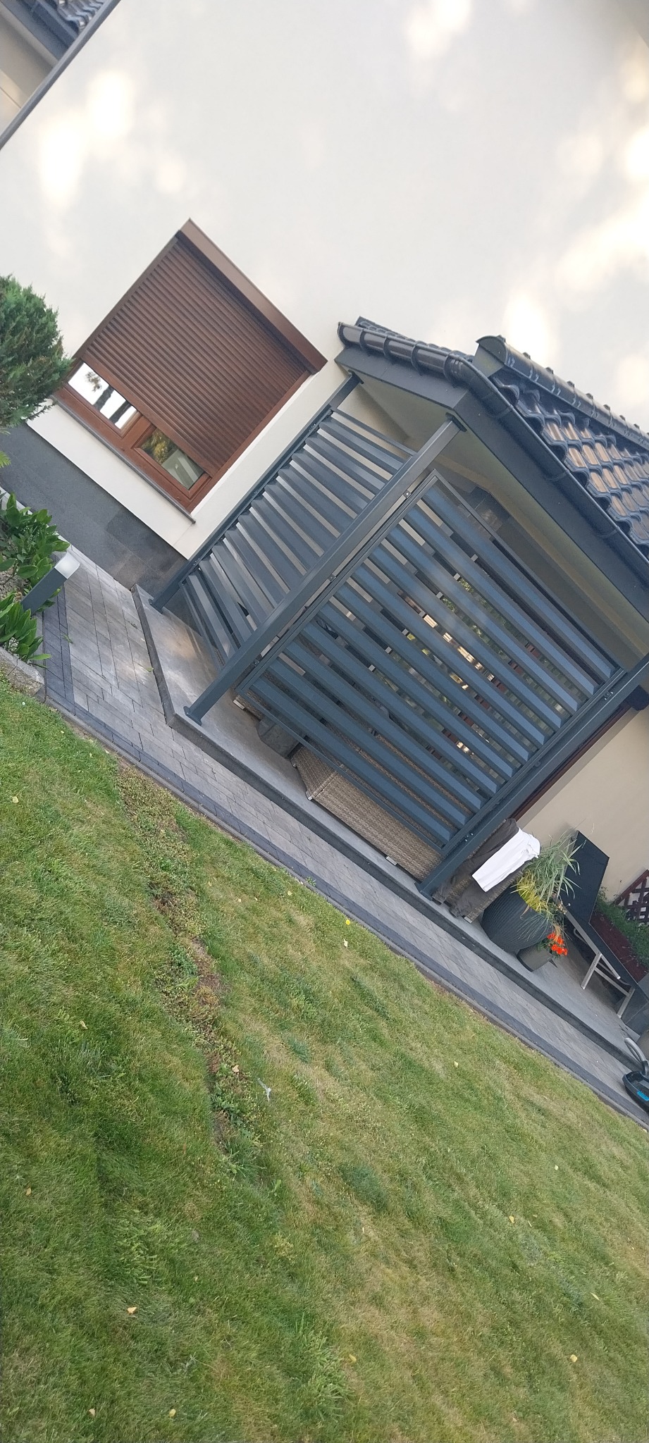 Nowoczesna pergola z szarymi, poziomymi lamelami, przylegająca do domu z brązowymi roletami w oknie, widok z trawnika.