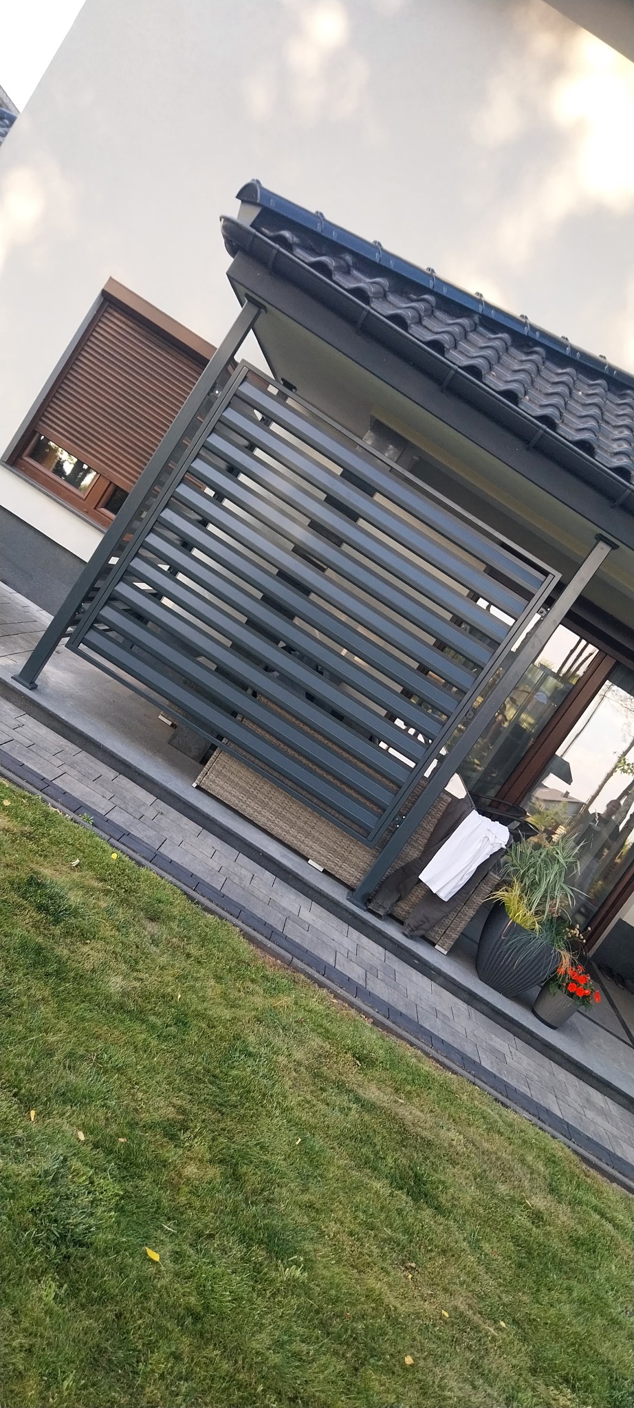Nowoczesna, szara konstrukcja z poziomych profili, prawdopodobnie zadaszenie tarasu, w Korytnicy. W tle widoczny dom z oknem i roletami zewnętrznymi.