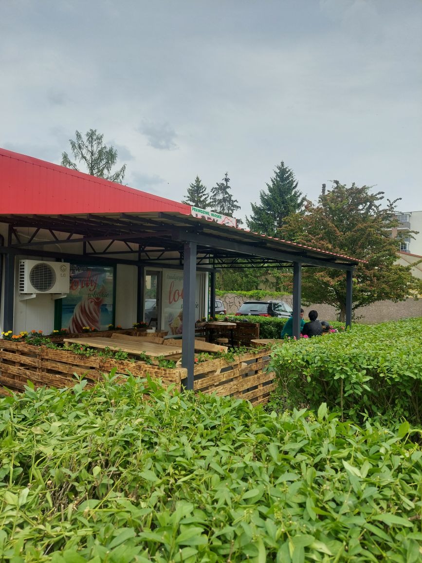 Zadaszona konstrukcja z metalowymi słupami i drewnianym wykończeniem, prawdopodobnie taras kawiarni, z widocznymi paletami tworzącymi barierki i podesty.