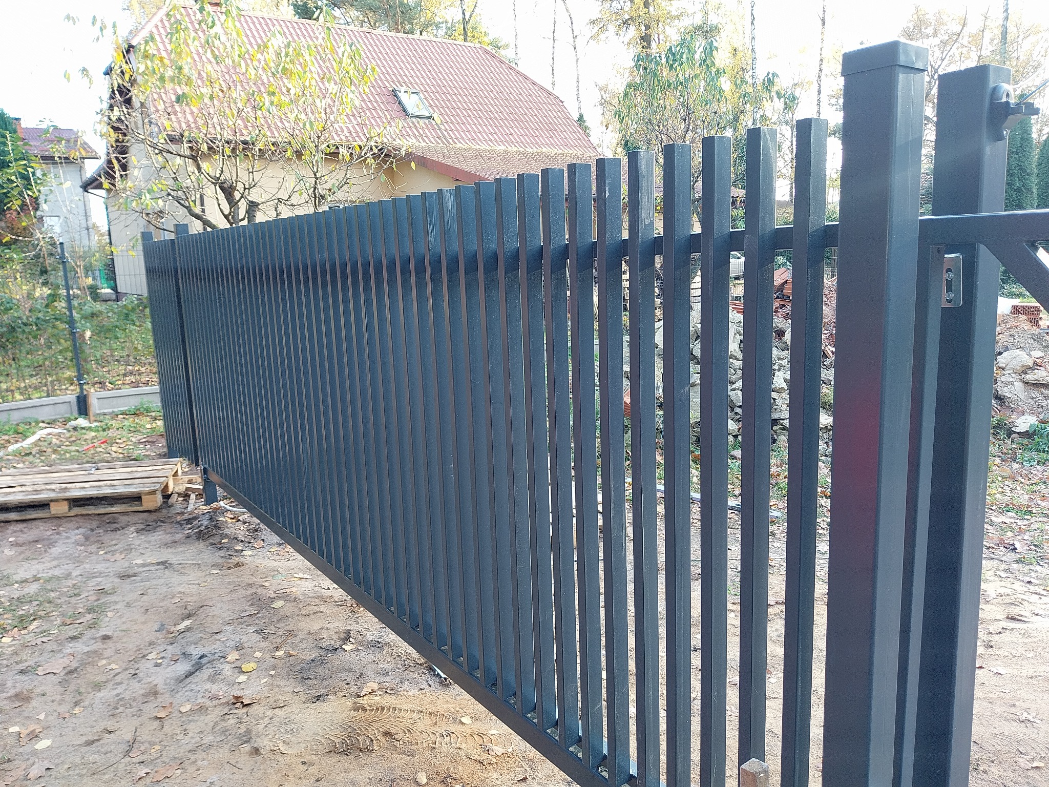 Nowoczesna, przesuwna brama wjazdowa w kolorze grafitowym, z pionowymi profilami, widoczna na tle domu z czerwoną dachówką i paletami na ziemi.