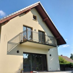 POL-MAT PAWEŁ PIOCHACZ - Dwupiętrowy dom z beżową elewacją, balkonem z czarną, metalową balustradą i brązowymi ramami okiennymi, sfotografowany w słoneczny dzień.