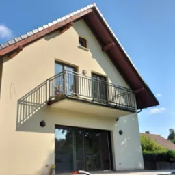 Dwupiętrowy dom z beżową elewacją, balkonem z czarną, metalową balustradą i brązowymi ramami okiennymi, sfotografowany w słoneczny dzień.