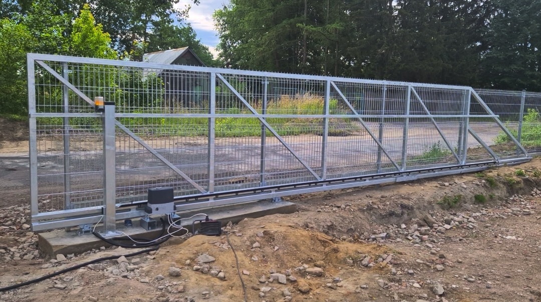 Długa, metalowa brama przesuwna z siatki panelowej, z automatyką i lampą ostrzegawczą, zamontowana na betonowym fundamencie przy drodze gruntowej.