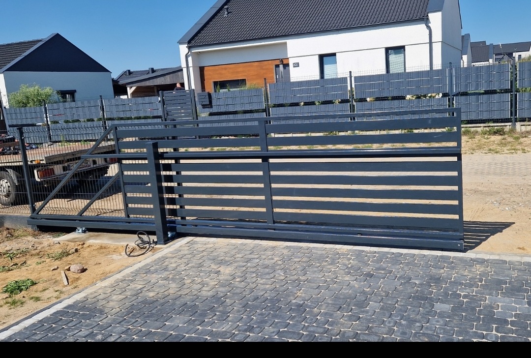 Szara, przesuwna brama wjazdowa z poziomymi szczeblami, widoczna na tle ogrodzenia panelowego i domów jednorodzinnych, zamontowana na kostce brukowej, z widoczną przyczepką transportową z tyłu.