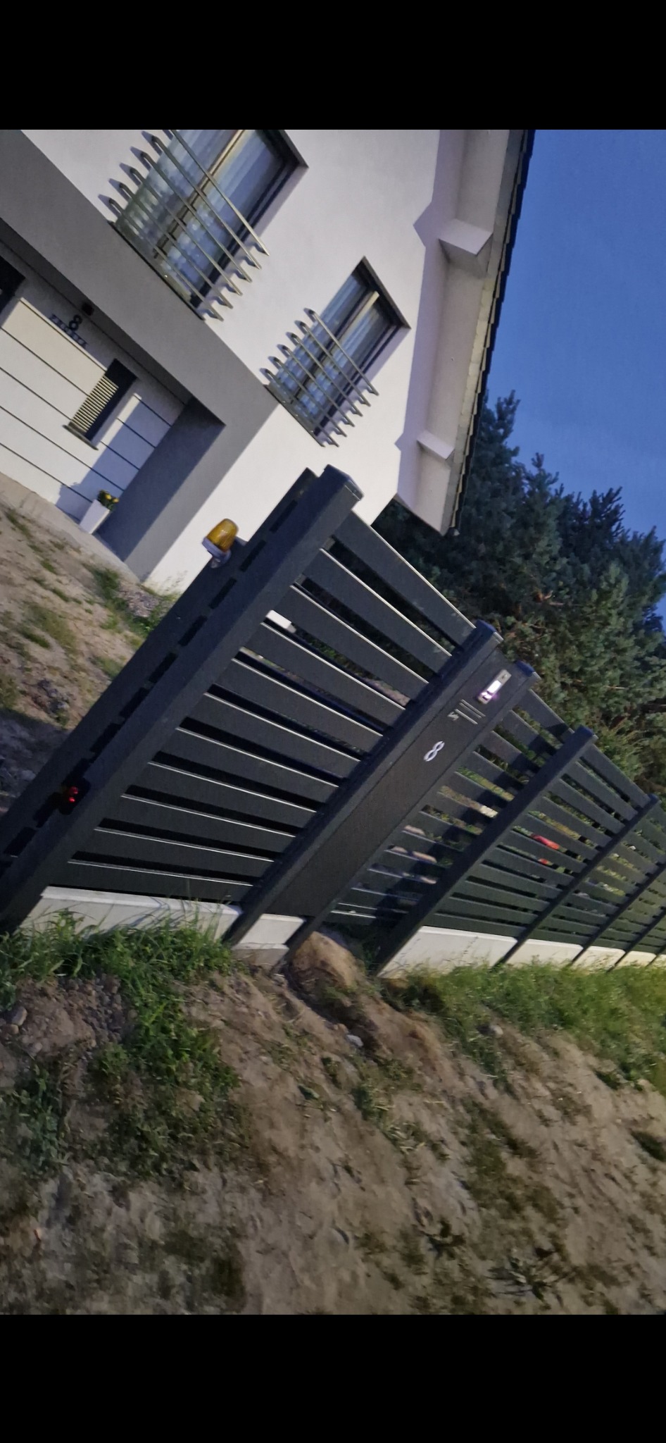 Nowoczesne, grafitowe ogrodzenie posesji z poziomych profili, widoczne z dołu pod kątem, z numerem domu '8' na słupku, na tle domu z białymi ścianami i szarymi elementami oraz ciemniejącego nieba.