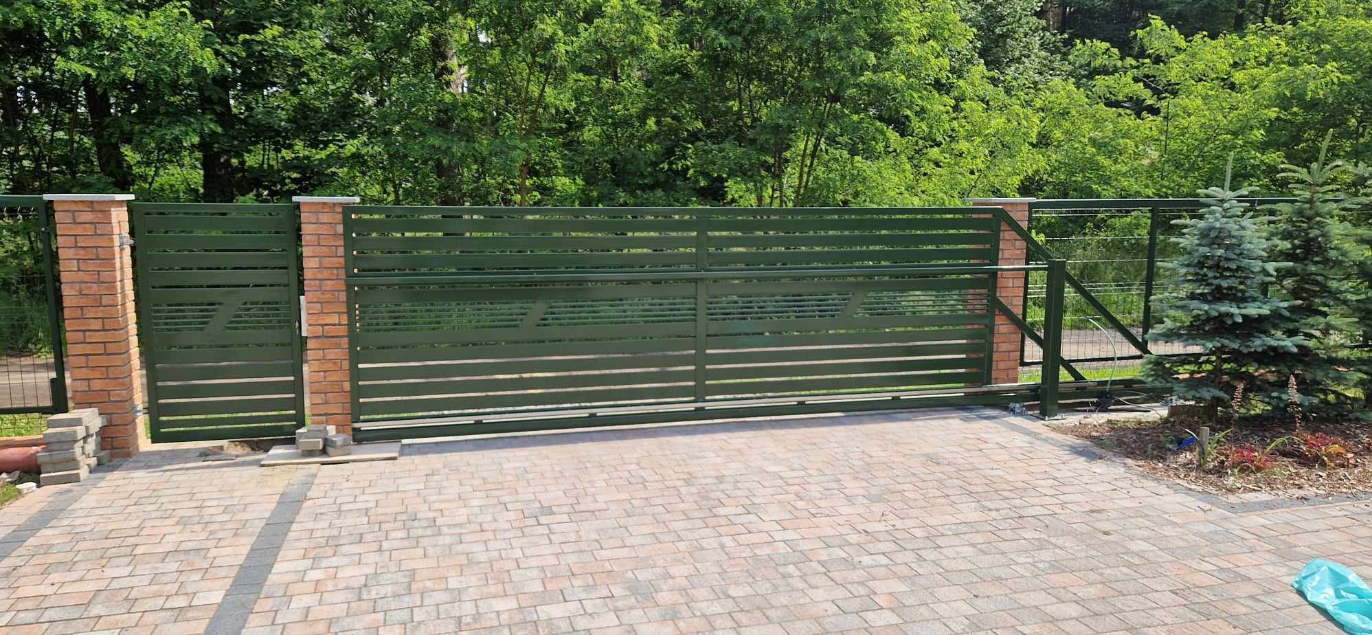 Zamykana, przesuwna brama wjazdowa z poziomych profili, pomalowana na oliwkowy kolor, osadzona pomiędzy ceglanymi słupkami na tle zieleni.