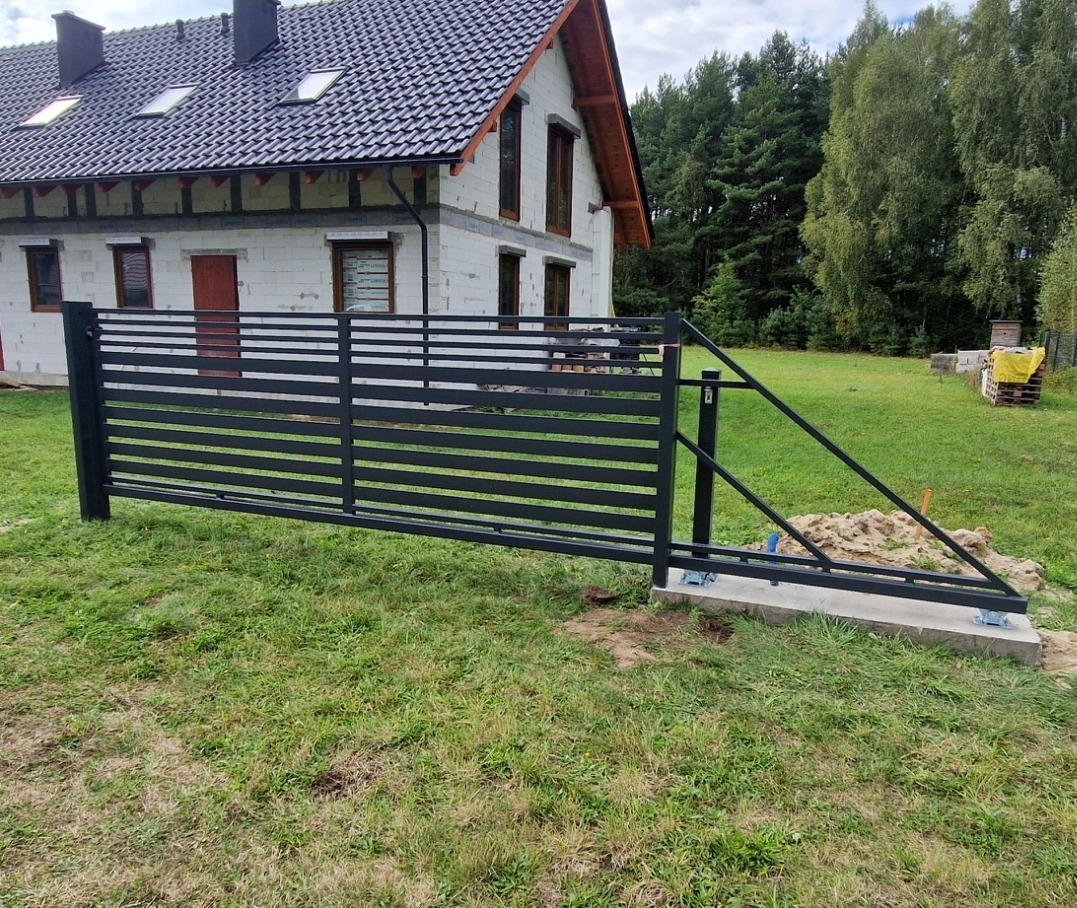 Czarna, pozioma brama wjazdowa z metalowych profili zamontowana na betonowej podstawie, na tle niedokończonego domu i zielonego trawnika.