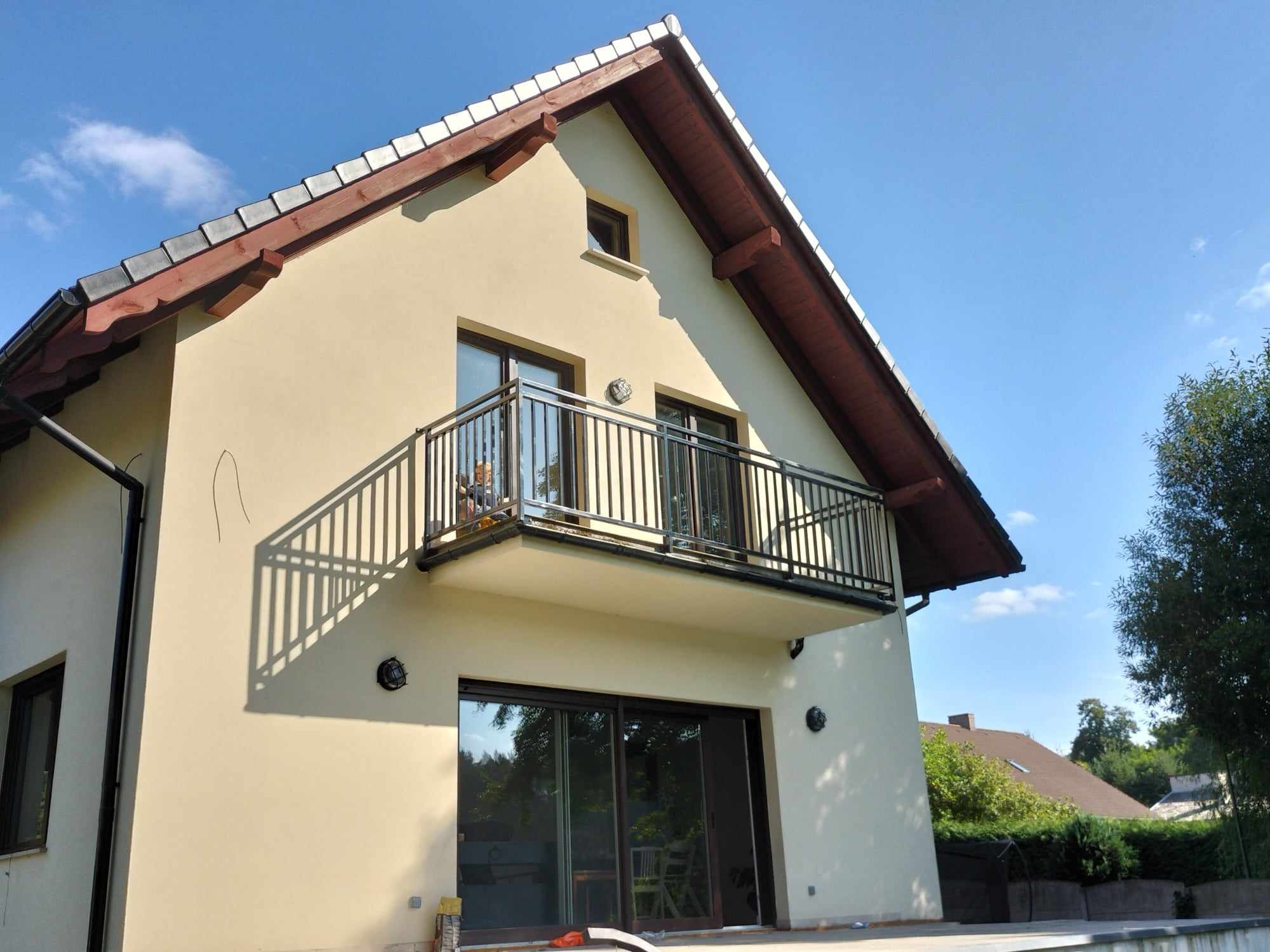 Dwupiętrowy dom z beżową elewacją, balkonem z czarną, metalową balustradą i brązowymi ramami okiennymi, sfotografowany w słoneczny dzień.
