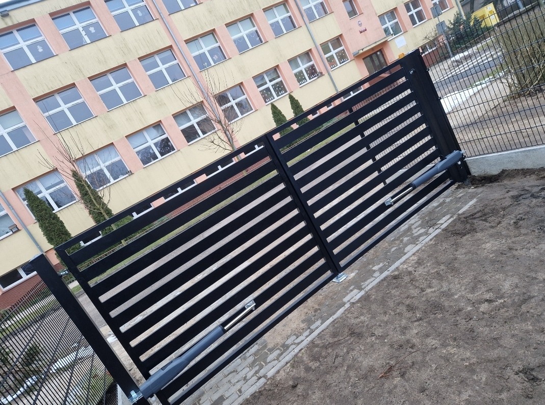 Czarna, pozioma brama wjazdowa z automatycznym napędem, widoczna na tle budynku szkolnego i ogrodzenia panelowego.