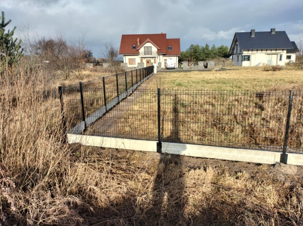 Nowoczesne ogrodzenie panelowe z podmurówką betonową, prowadzące wzdłuż ścieżki do domu w tle, widoczne trawy i roślinność w naturalnym otoczeniu.