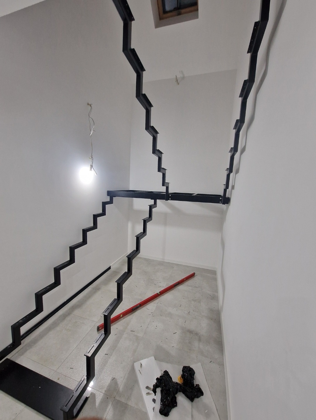 Wnętrze w trakcie montażu metalowej konstrukcji schodów z czarnych profili, widoczny poziomica i resztki pianki montażowej na podłodze.