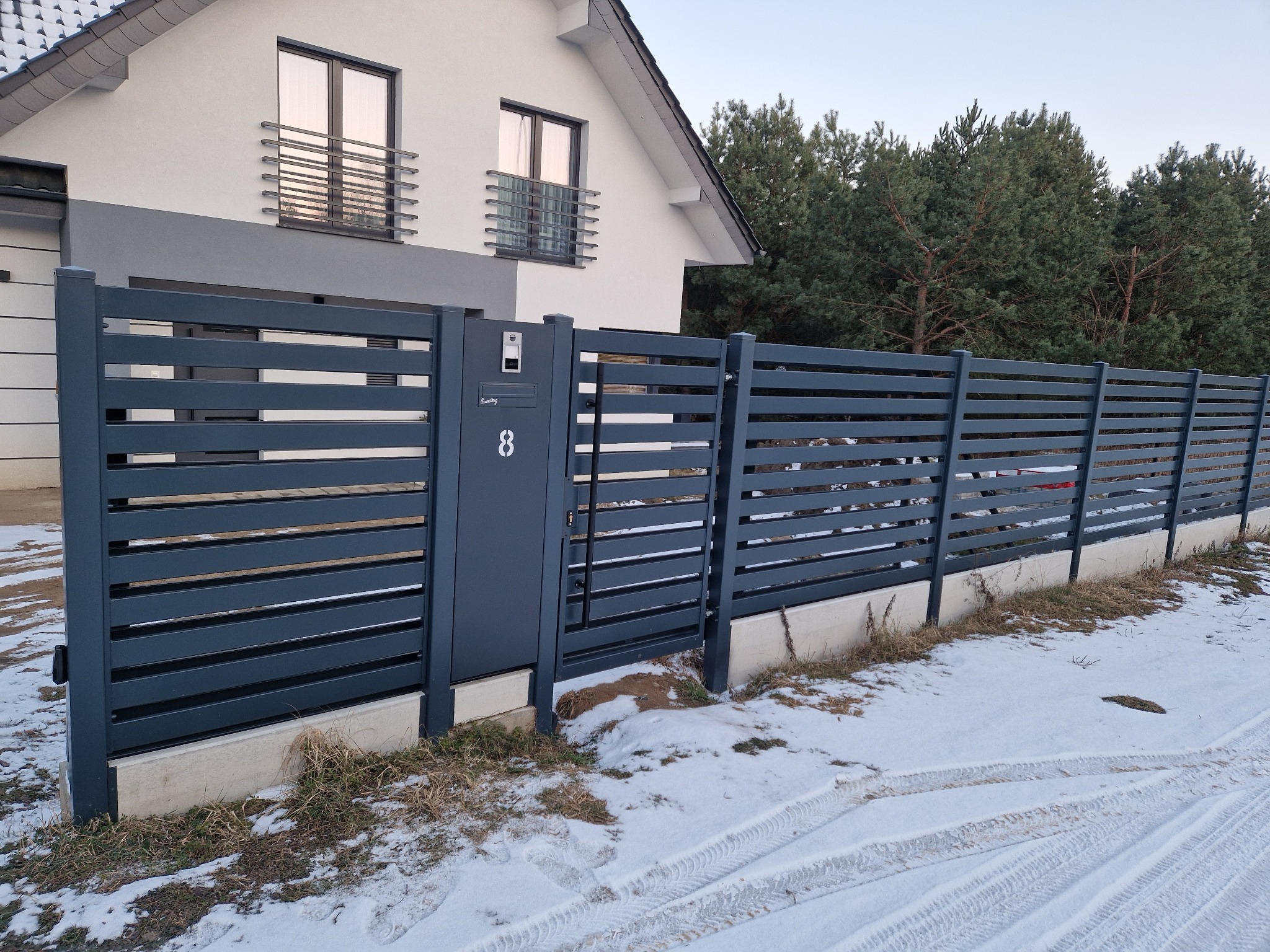 Nowoczesne, poziome ogrodzenie z szarych metalowych paneli z numerem domu '8' na skrzynce pocztowej, w zimowej scenerii z ośnieżonym trawnikiem i śladami opon.