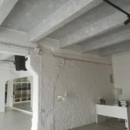 Jasne, przestronne wnętrze z betonowym sufitem, oświetleniem szynowym i białą ceglaną ścianą z widocznymi instalacjami, uzupełnione minimalistycznym biurkiem z lampą i książkami.