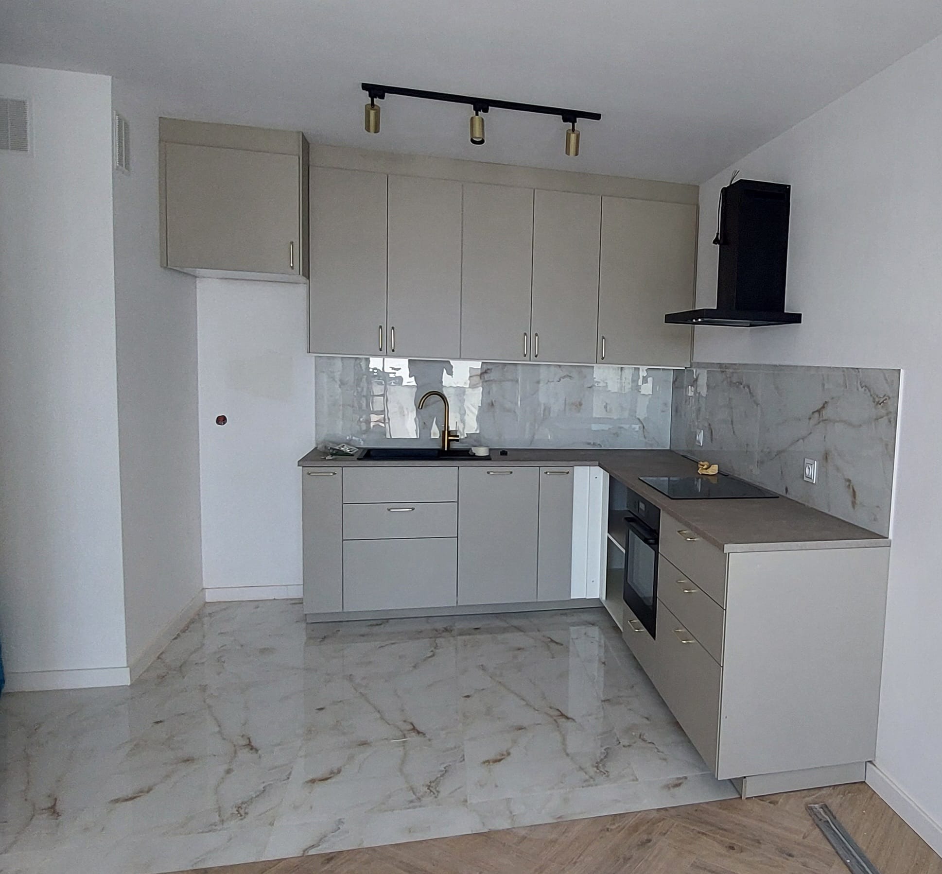 Jasna kuchnia z marmurowym blatem i podłogą, oświetlona reflektorami. Nowoczesny design z czarnym okapem i złotą armaturą. Widok na wykończone wnętrze.