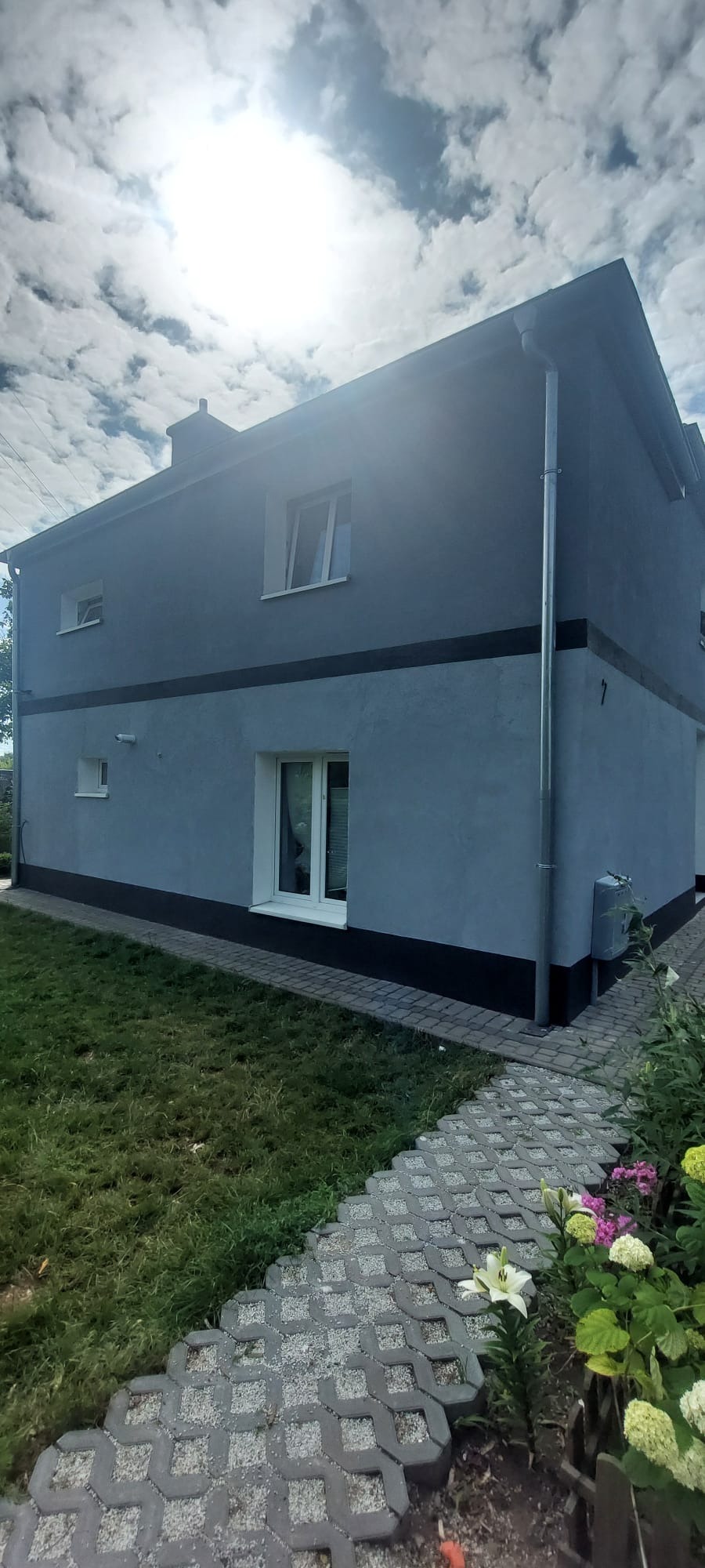 Dwukondygnacyjny dom z szarą elewacją, czarnym pasem i białymi oknami, widziany z poziomu trawnika i ścieżki z ażurowych płyt betonowych w słoneczny dzień.