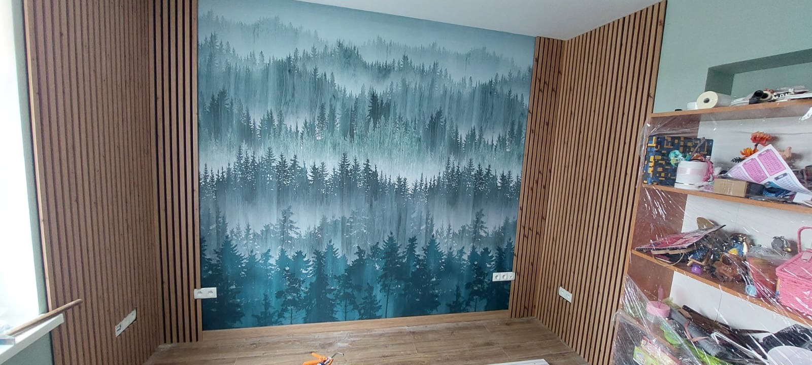 Pokój po wykończeniu: ściana z fototapetą leśną, obłożona drewnianymi lamelami. Widoczne gniazdka elektryczne i fragment regału z zabezpieczeniem folią malarską.