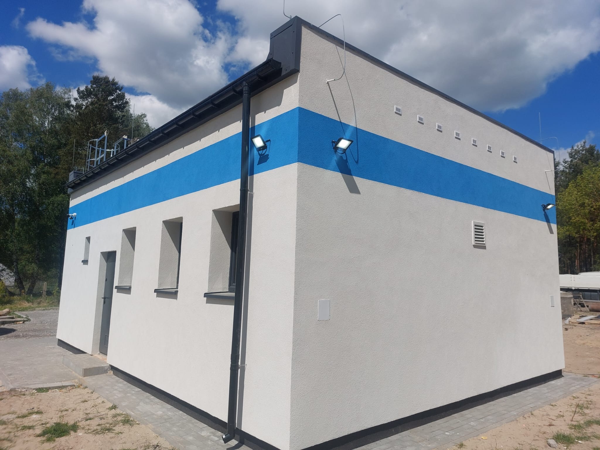 Nowoczesny budynek z szarą elewacją i niebieskim pasem, oświetlony reflektorami, z widocznymi oknami i drzwiami, na tle błękitnego nieba z chmurami.