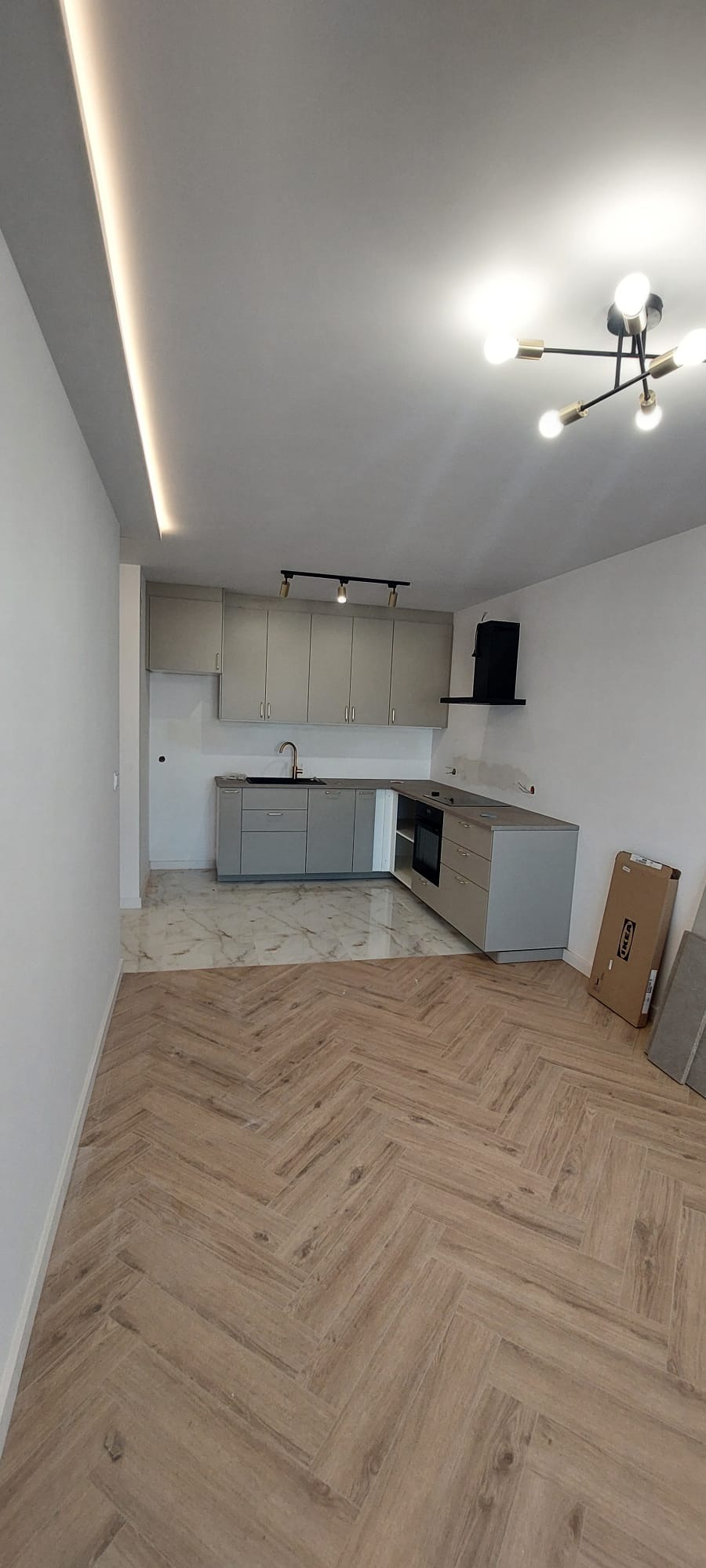 Jasna kuchnia z szarymi szafkami, marmurową podłogą i drewnianą podłogą ułożoną w jodełkę. Widoczne oświetlenie sufitowe i karton IKEA.