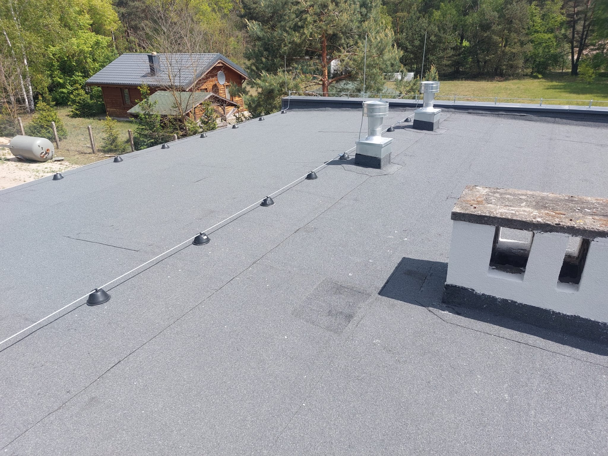 Szary dach pokryty papą termozgrzewalną z kominami wentylacyjnymi i systemem odgromowym na tle drewnianego domu i zieleni.