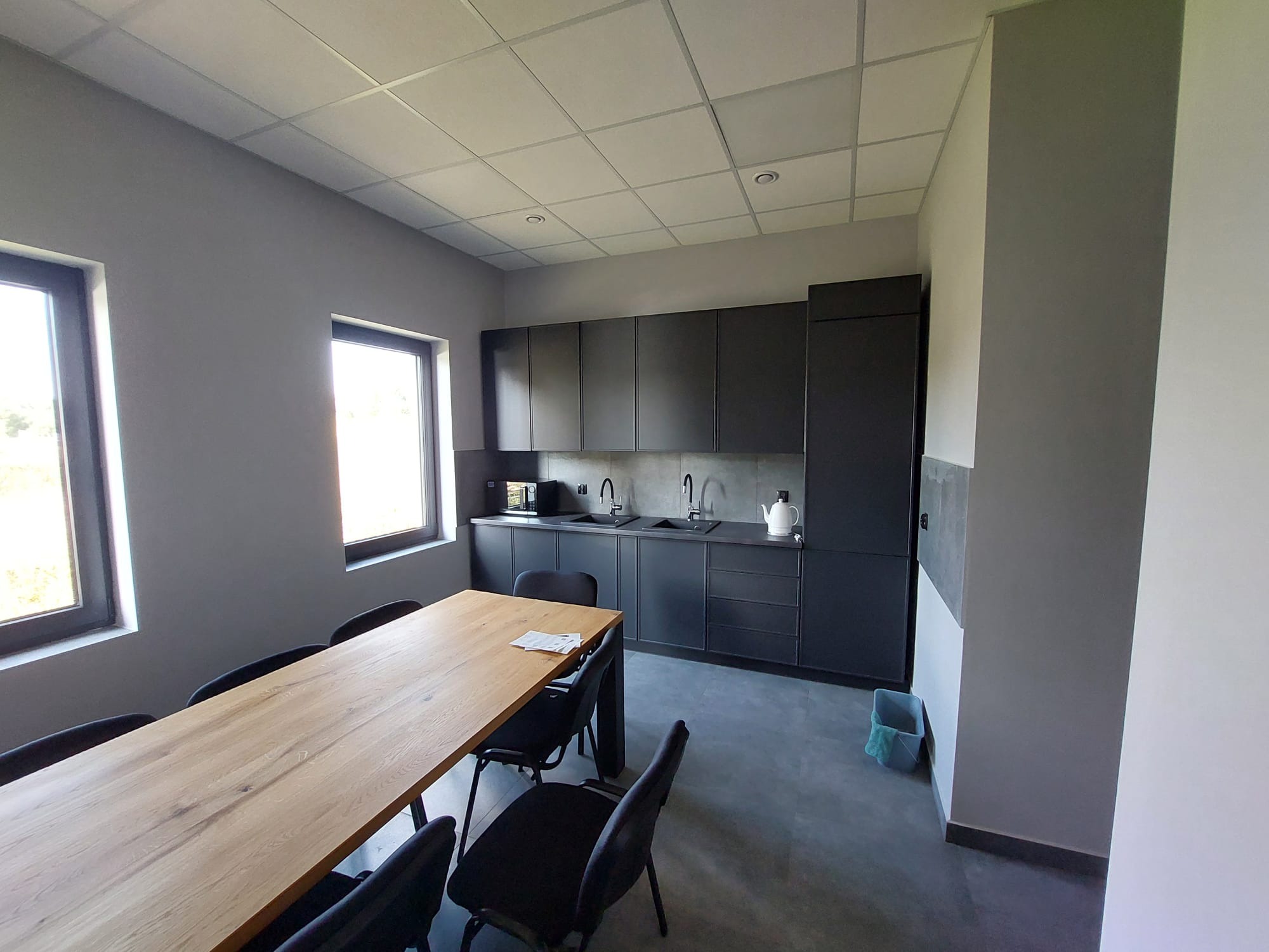 Nowoczesna, szara kuchnia biurowa z drewnianym stołem i czarnymi krzesłami. Minimalistyczny design, dwa okna, oświetlenie sufitowe.