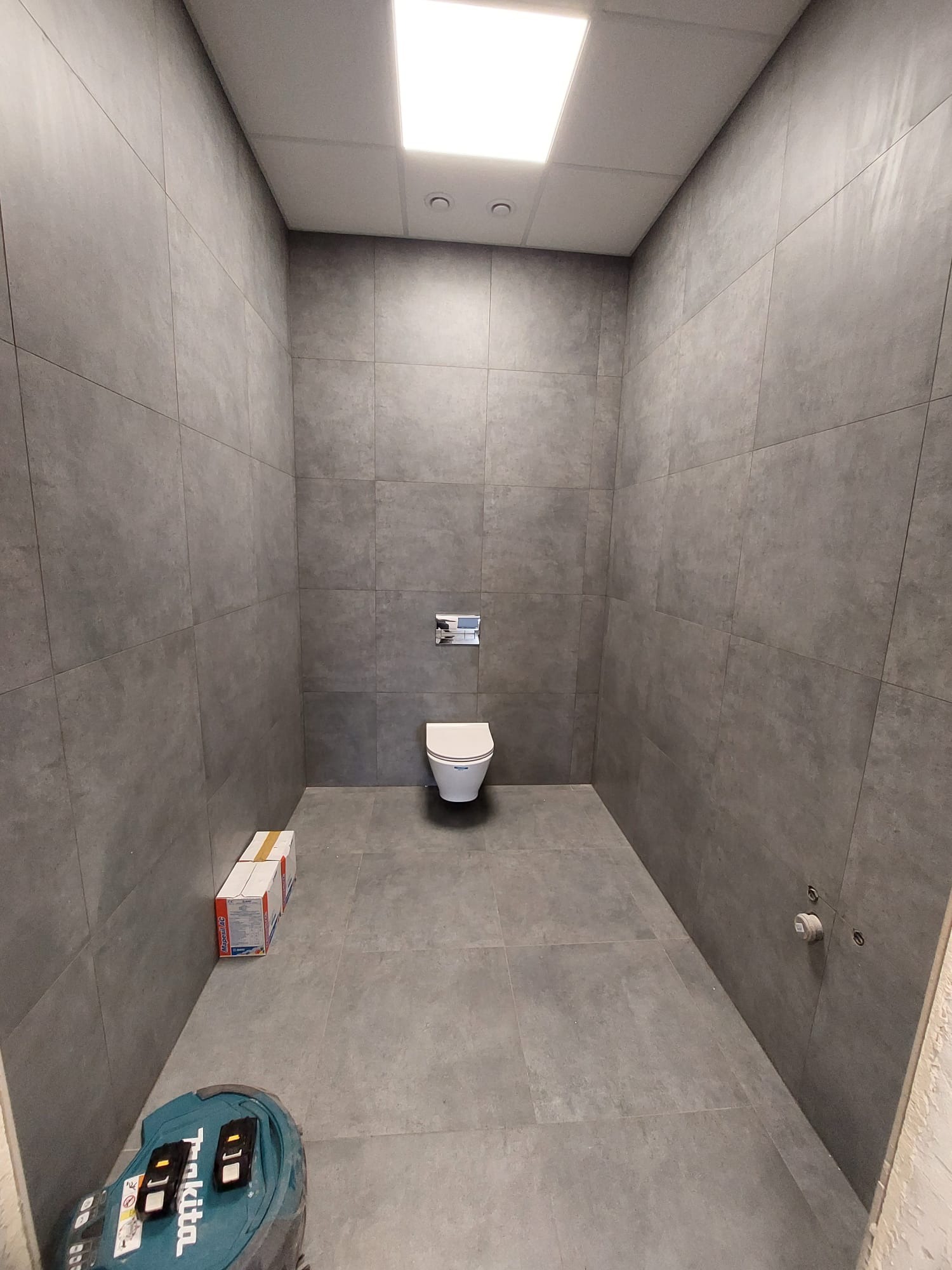 Wykończona łazienka w szarościach z wiszącą toaletą. Widoczne płytki na ścianach i podłodze, pudełko z materiałami i narzędzia. Minimalistyczny design.