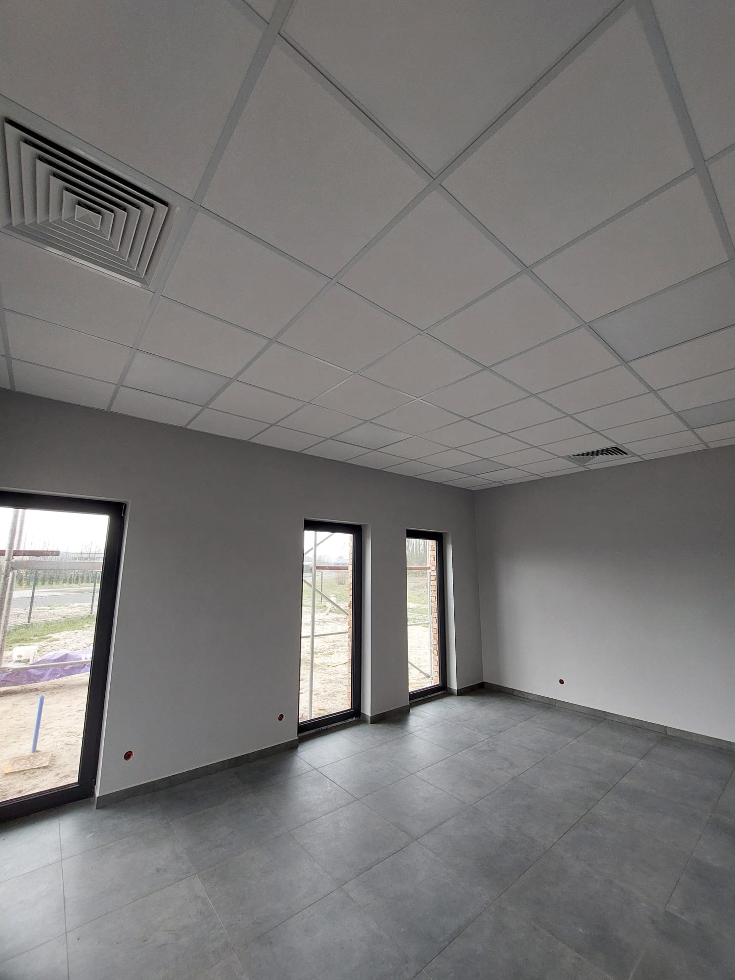Szare płytki podłogowe i sufit podwieszany kasetonowy z kratką wentylacyjną w nowym, pustym pomieszczeniu z trzema oknami.