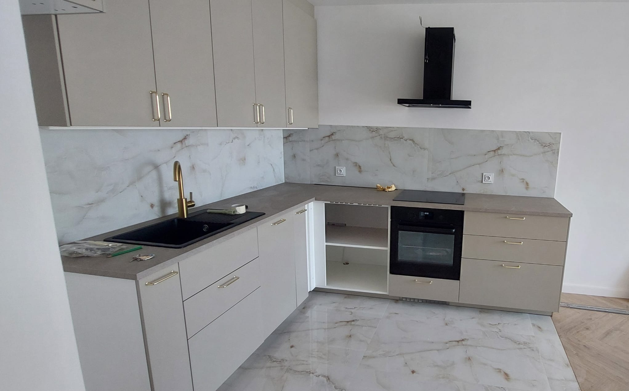 Jasna kuchnia z marmurowym blatem i złotą armaturą w Bydgoszczy. Nowoczesne szafki wiszące i dolne, czarny okap. Płytki imitujące marmur na ścianie.