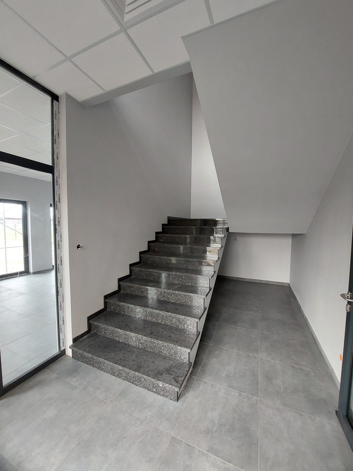Wnętrze z szarymi, granitowymi schodami i podłogą z płytek. Widoczne drzwi z czarną ramą i przeszklona ściana. Minimalistyczny design, surowe wykończenie.