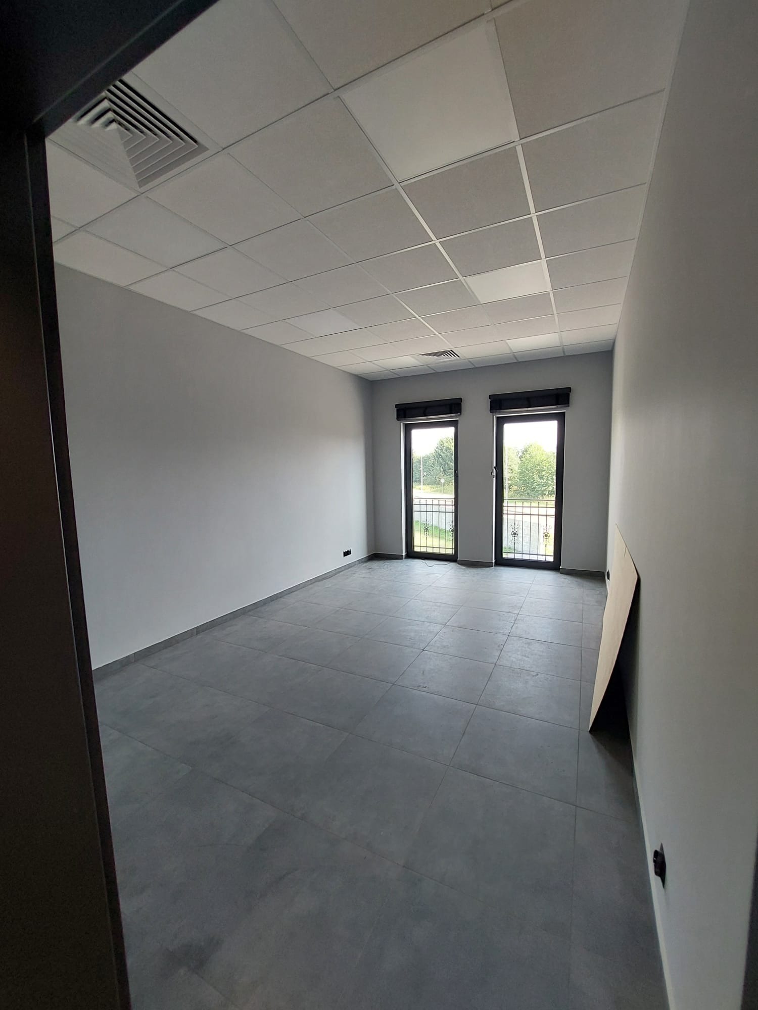 Szare płytki podłogowe w pustym, jasnym pomieszczeniu biurowym z dwoma oknami balkonowymi i podwieszanym sufitem kasetonowym.
