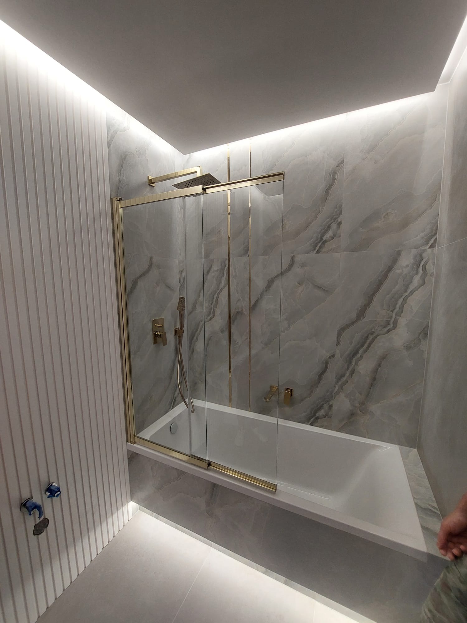 Nowoczesna łazienka z wanną i szklanym parawanem w złotej ramie, marmurowe płytki na ścianach, oświetlenie LED, minimalistyczny design.