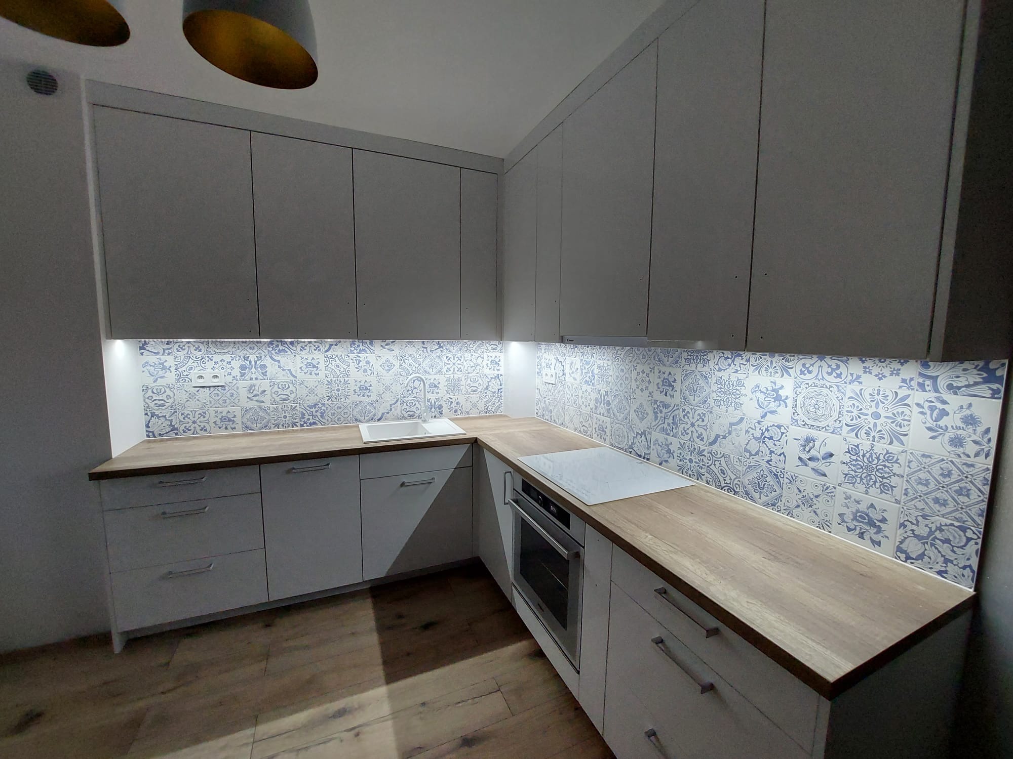 Jasna kuchnia z drewnianym blatem i dekoracyjnymi, niebiesko-białymi płytkami nad blatem. Widoczne białe szafki i sprzęt AGD.