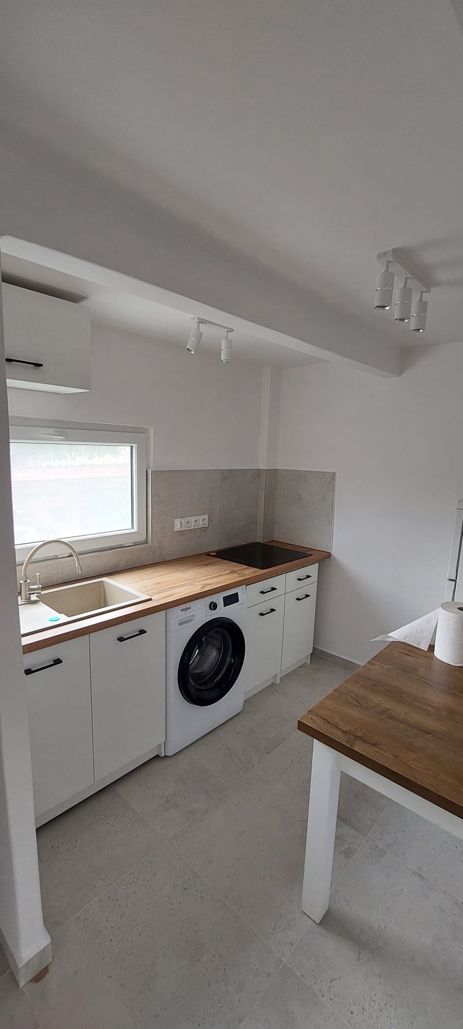Jasna kuchnia z drewnianym blatem, białymi szafkami i pralką w zabudowie. Minimalistyczne oświetlenie punktowe i szara podłoga tworzą nowoczesny klimat.
